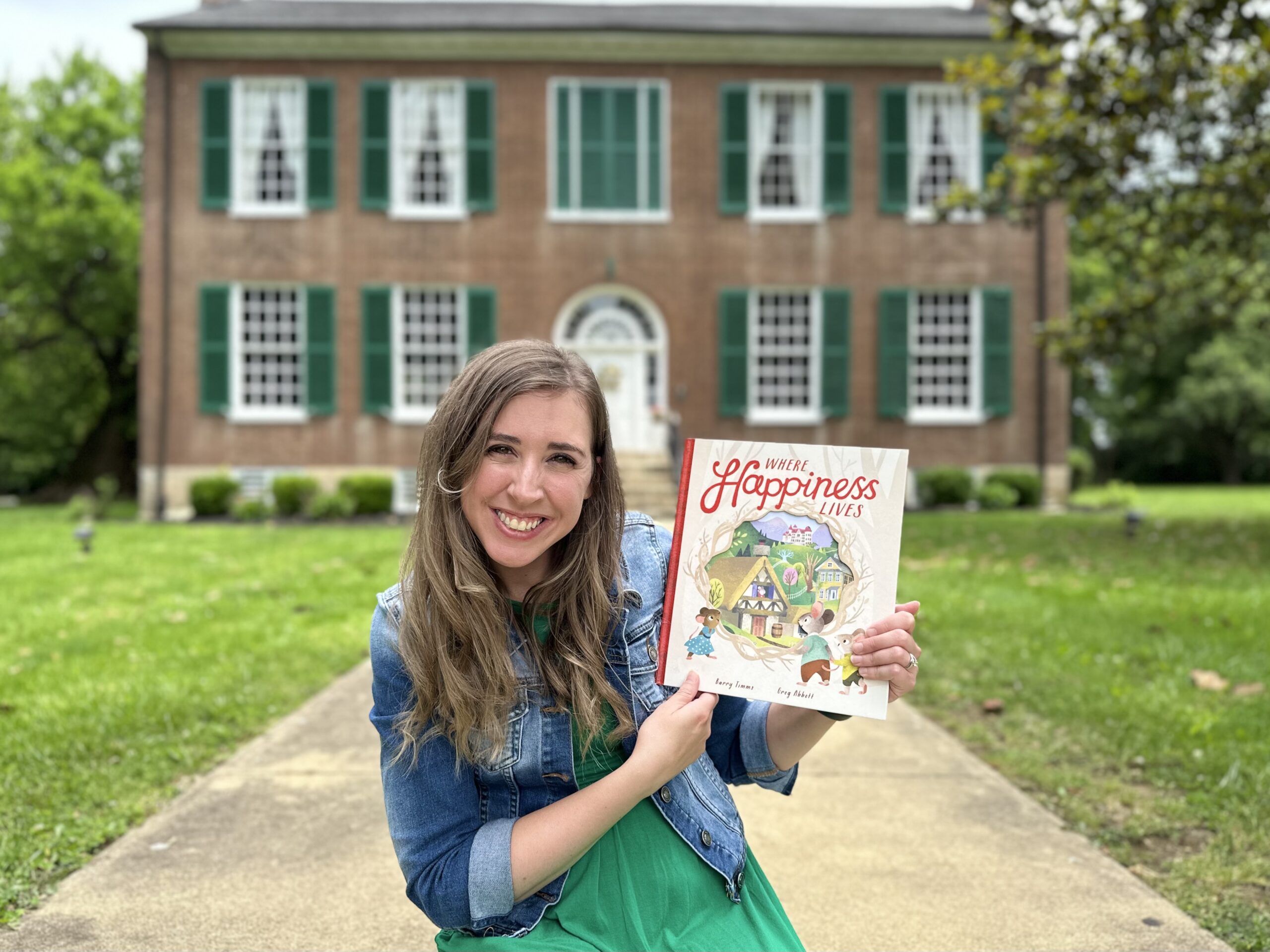Jen Williams holding a children's book, smiling in front of a teal book backdrop